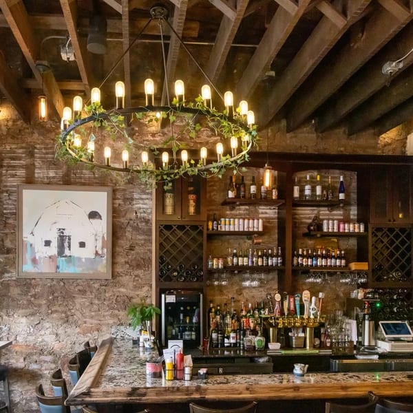 Rustic bar interior with exposed brick, wooden beams, a large chandelier, and a fully stocked bar.