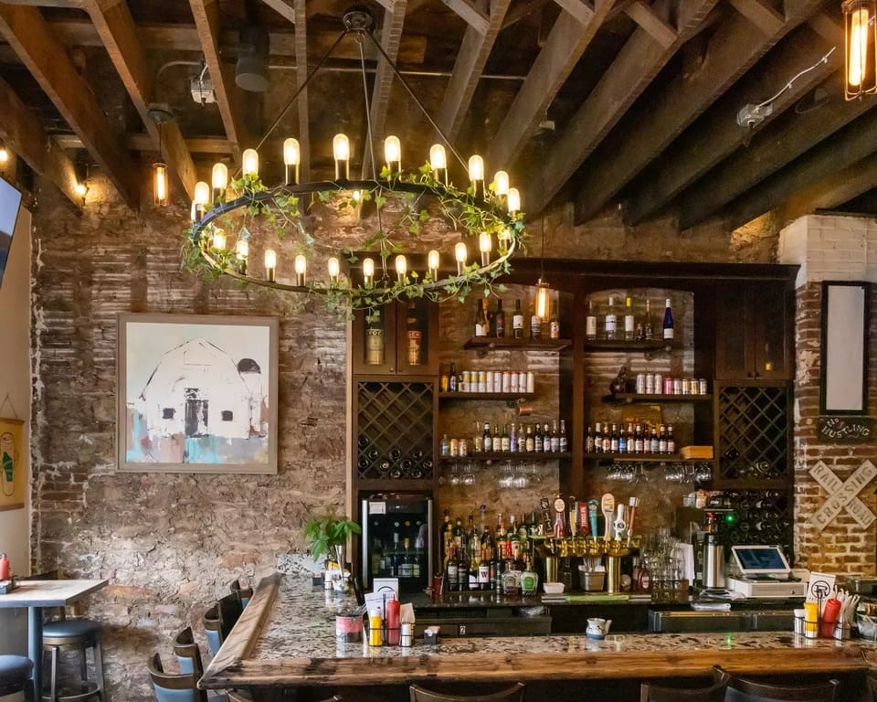Rustic bar interior with exposed brick, wooden beams, a large chandelier, and a fully stocked bar.