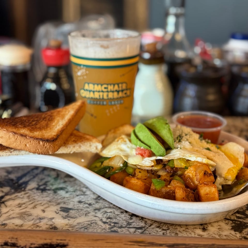 A breakfast skillet with eggs, avocado, potatoes, and toast, next to a beer.