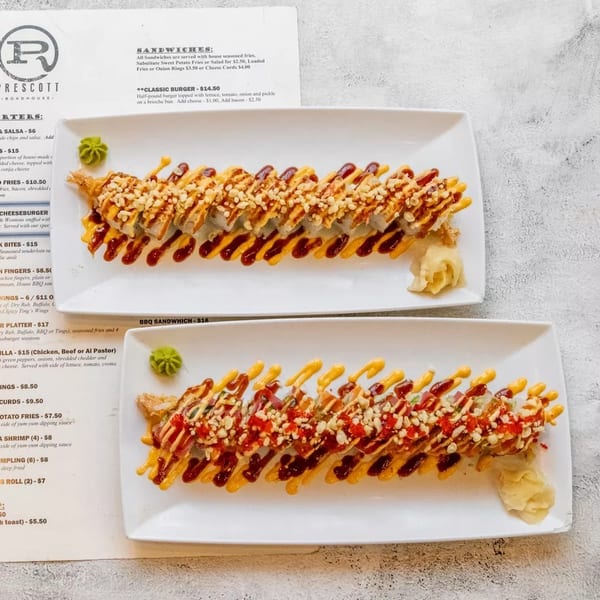 Two plates of Prescott Roadhouse sushi rolls with sauces, next to a menu on a gray surface.