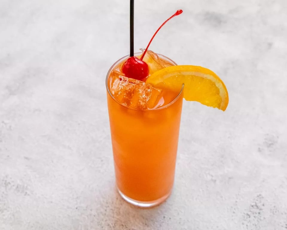 Orange cocktail with ice, garnished with a cherry, orange slice, and black straw.