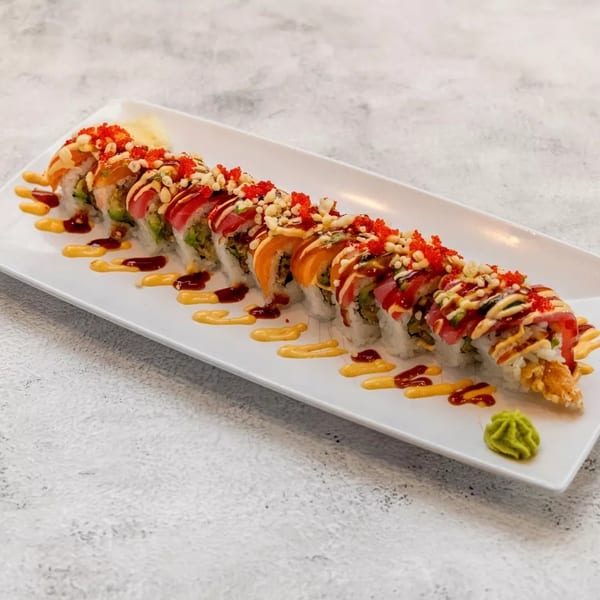 Long sushi roll with various fish, sauces, and toppings on a white rectangular plate.