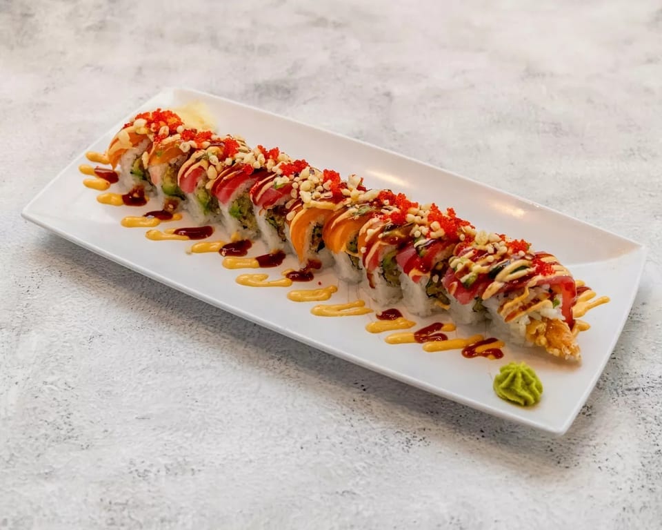 Long sushi roll with various fish, sauces, and toppings on a white rectangular plate.
