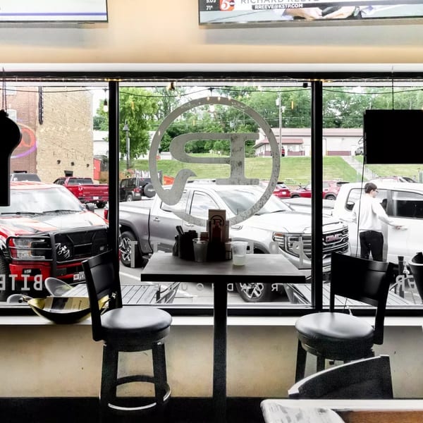 Interior view of a restaurant with high-top tables and chairs, looking out a window at a parking lot.