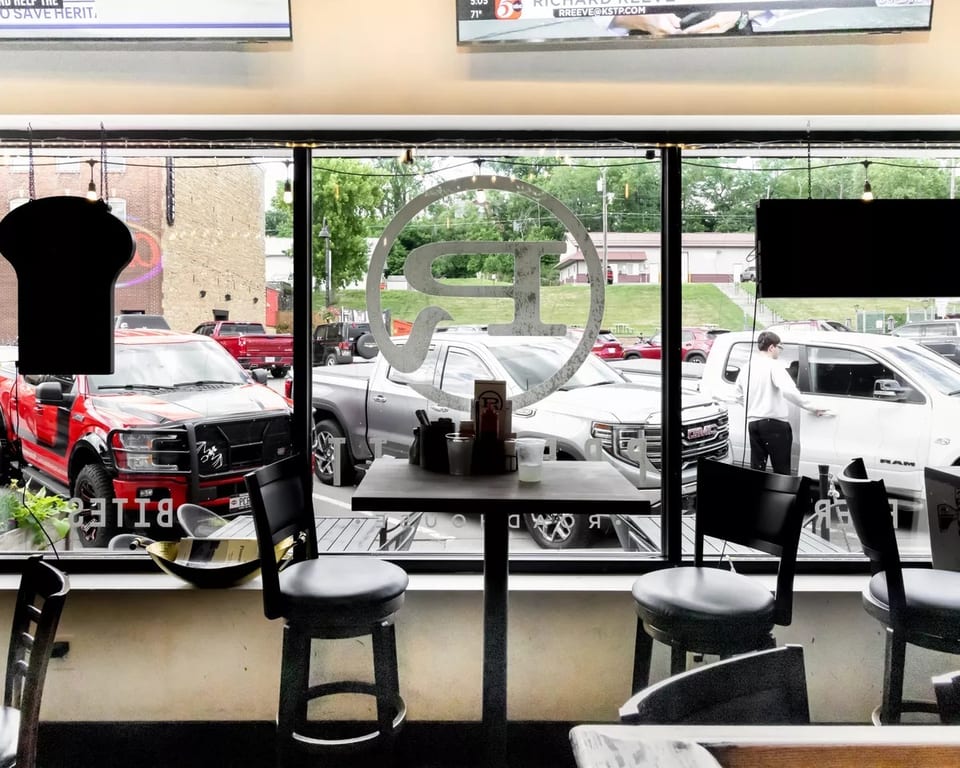 Interior view of a restaurant with high-top tables and chairs, looking out a window at a parking lot.