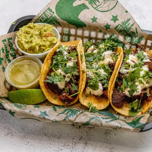 Three tacos with meat, cilantro, and sauce, served with guacamole, salsa, and lime.