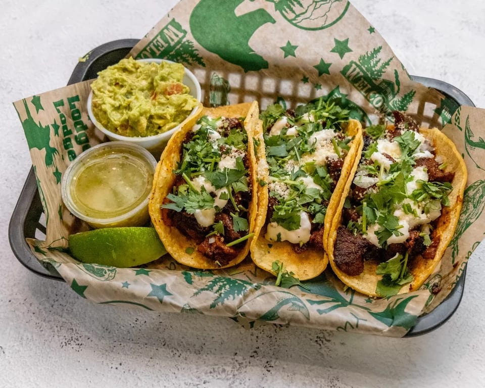 Three tacos with meat, cilantro, and sauce, served with guacamole, salsa, and lime.